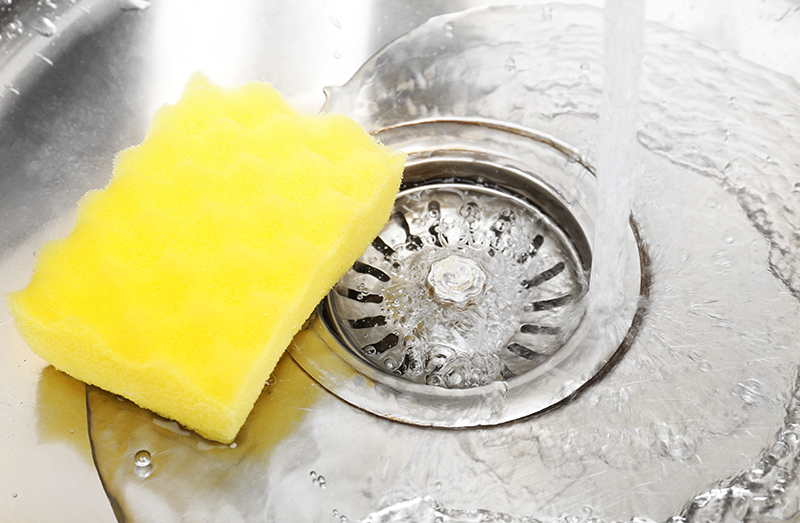 A stainless steel sink with water going down the drain and a yellow sponge on top.